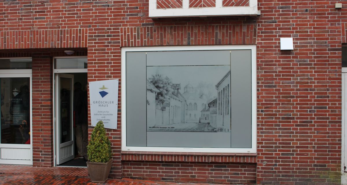 Die Fassade des GröschlerHauses, Zentrum für Jüdische Geschichte und Zeitgeschichte der Region. Das Gebäude ist ein Rotklinkerbau mit Schaufenstern, neben der Eingangstür hängt der Namensschriftzug der Institution auf einem weißen Schild. Im Schaufenster daneben ist eine historische Abbildung der inzwischen zerstörten jeverschen Synagoge zu sehen.